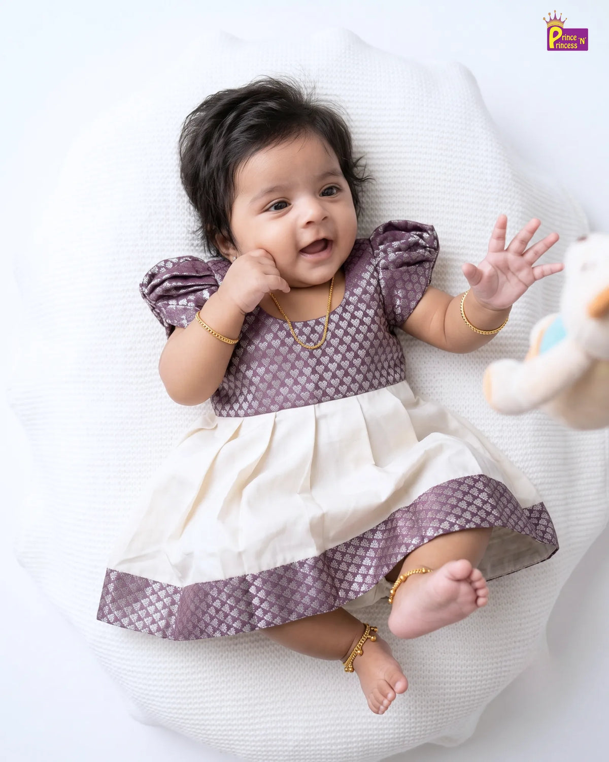 A baby girl lying on a white textured cushion, wearing a two-tone dress with a purple patterned bodice and skirt border, and an off-white skirt. She is accessorized with gold jewelry, and a plush toy is visible on the right side.