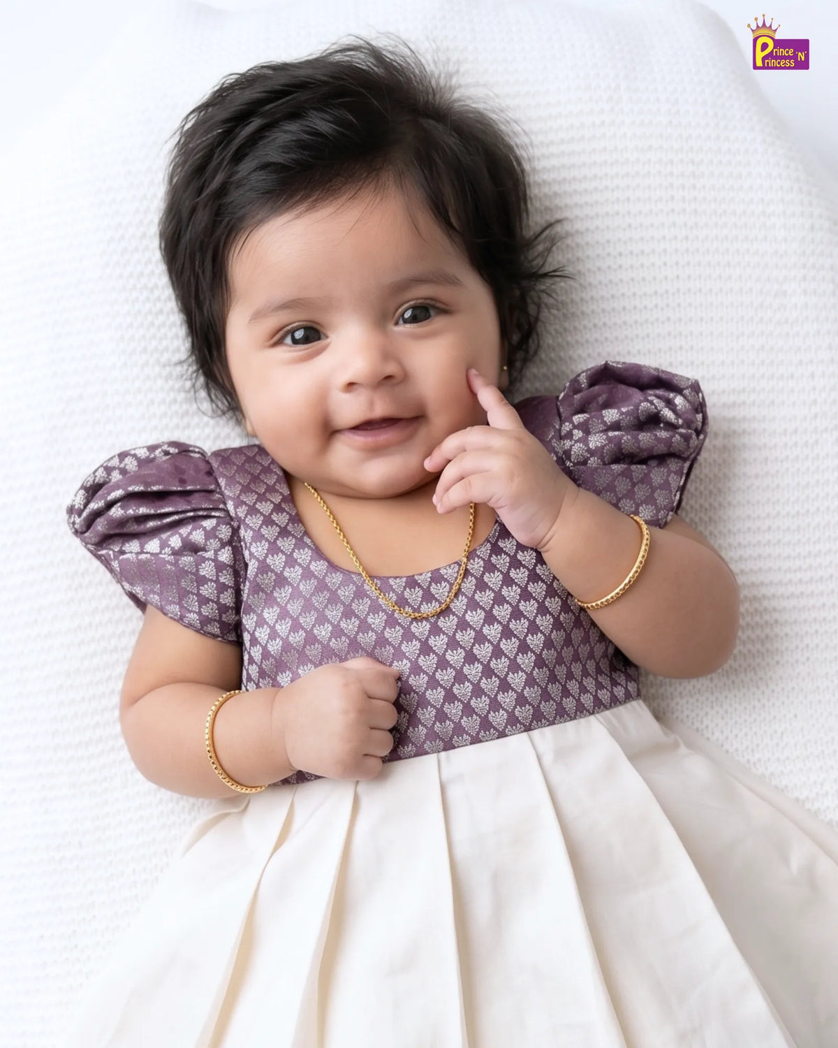 A baby girl lying on a white textured blanket, wearing a purple and white patterned top with a cream-colored skirt, along with gold jewelry.