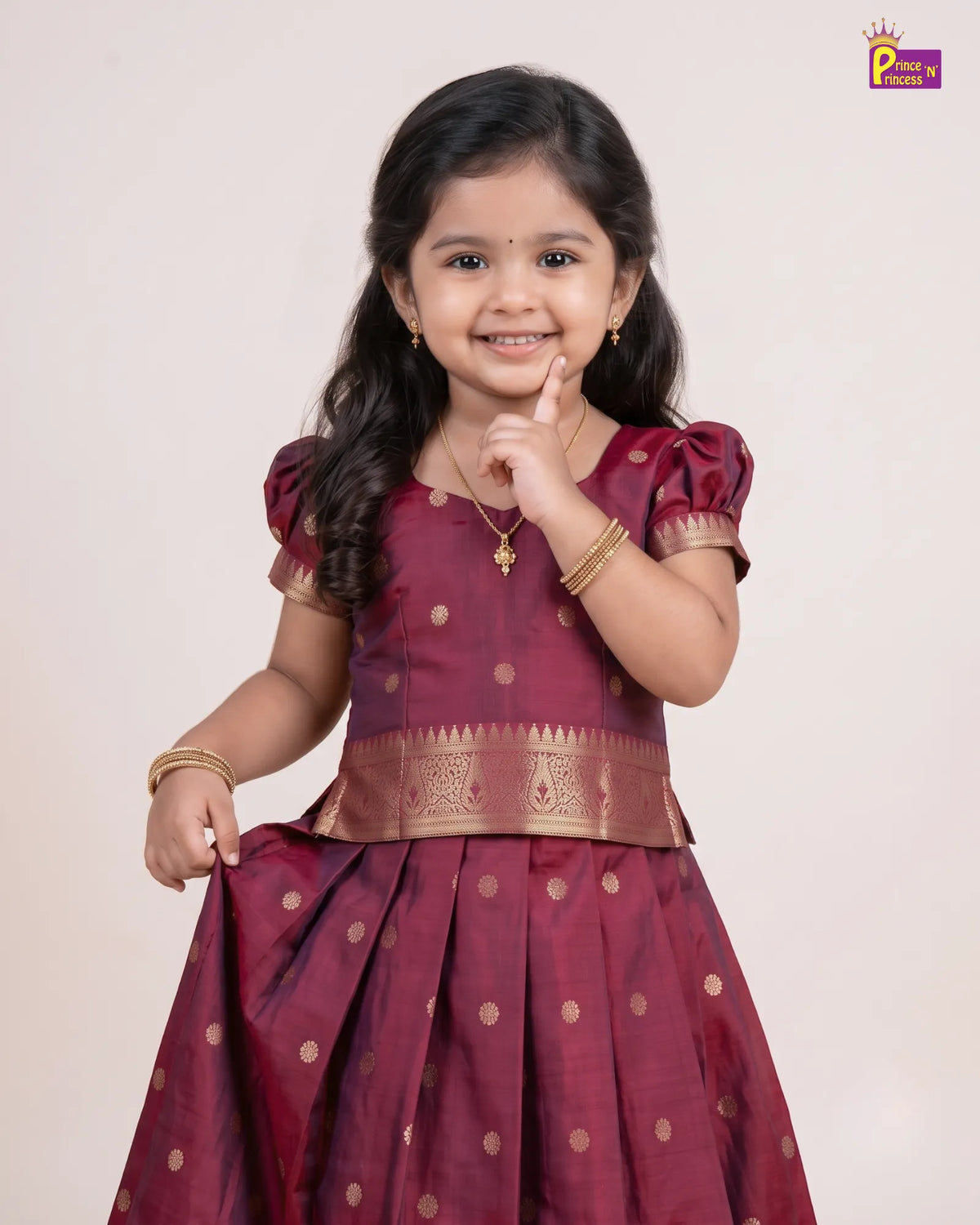 A young girl modeling a traditional Indian dress, featuring a deep maroon skirt with gold polka dots and a matching maroon and gold embroidered bodice, set against a plain light beige background.