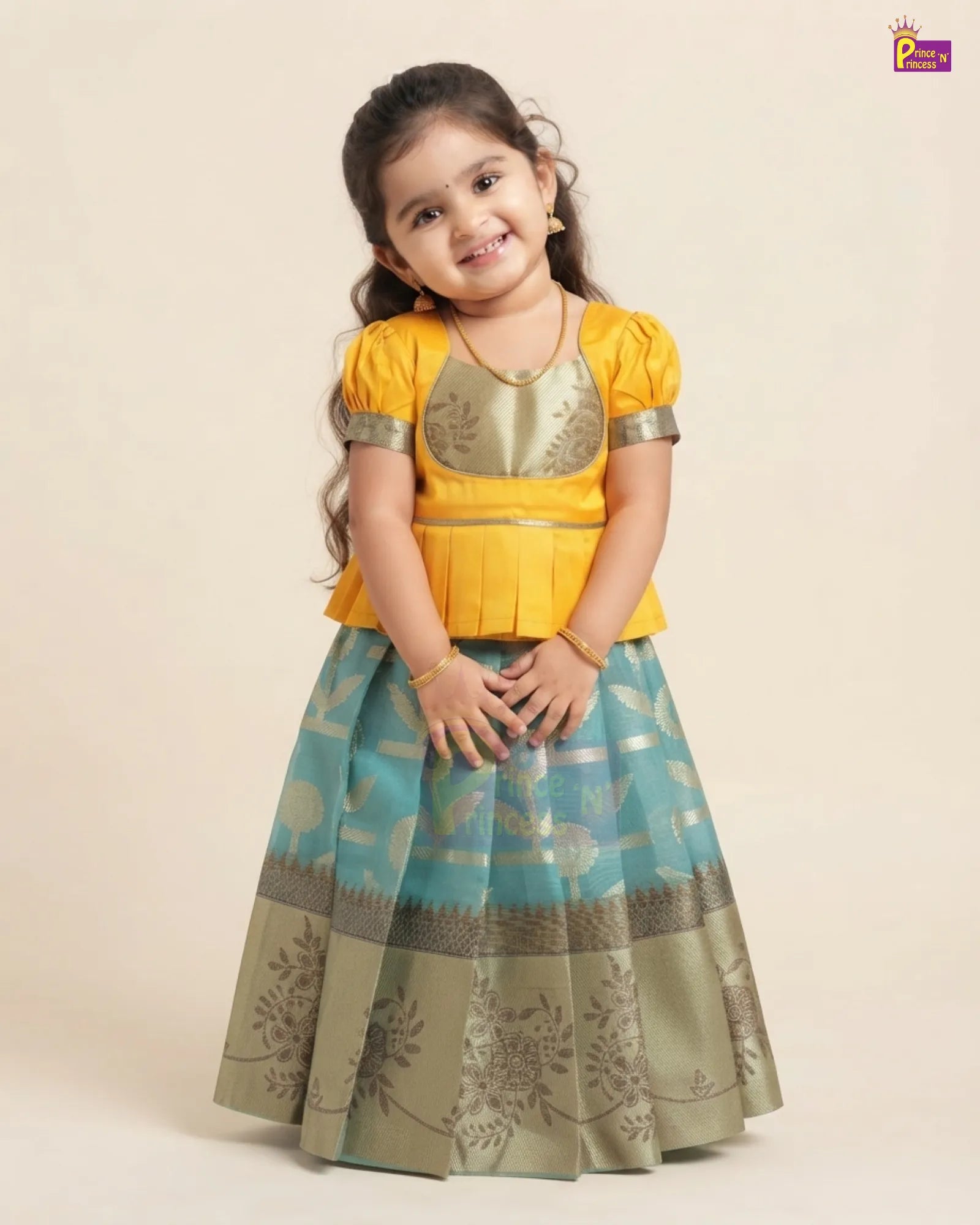 A young girl modeling a traditional Indian outfit consisting of a bright yellow short-sleeve blouse with metallic gold embroidery and a flared teal blue skirt with gold floral patterns and a metallic olive-gold border, set against a plain beige background.