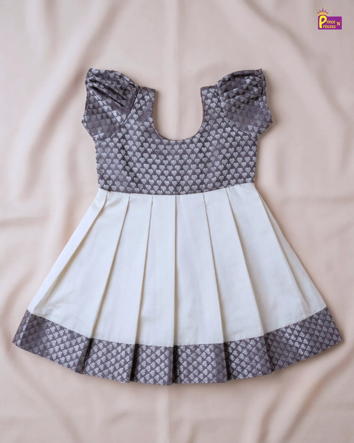 A baby girl's dress with a gray and white patterned bodice and hem, and a solid white pleated skirt, laid flat on a beige fabric background.