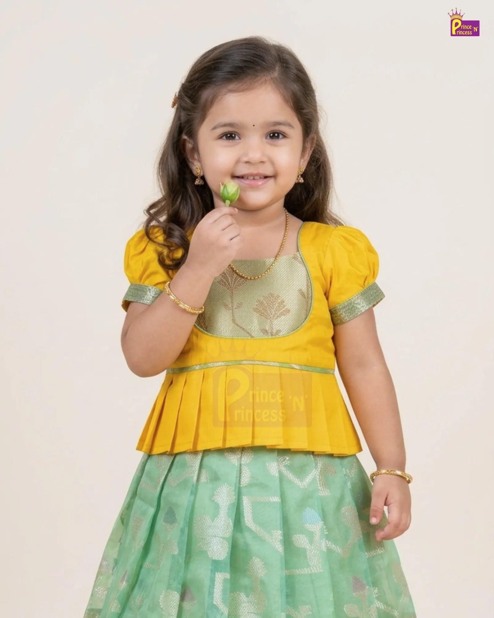 A young girl modeling a traditional Indian outfit consisting of a yellow top with green trim and a light green patterned skirt, holding a small green leaf against a plain light beige background.