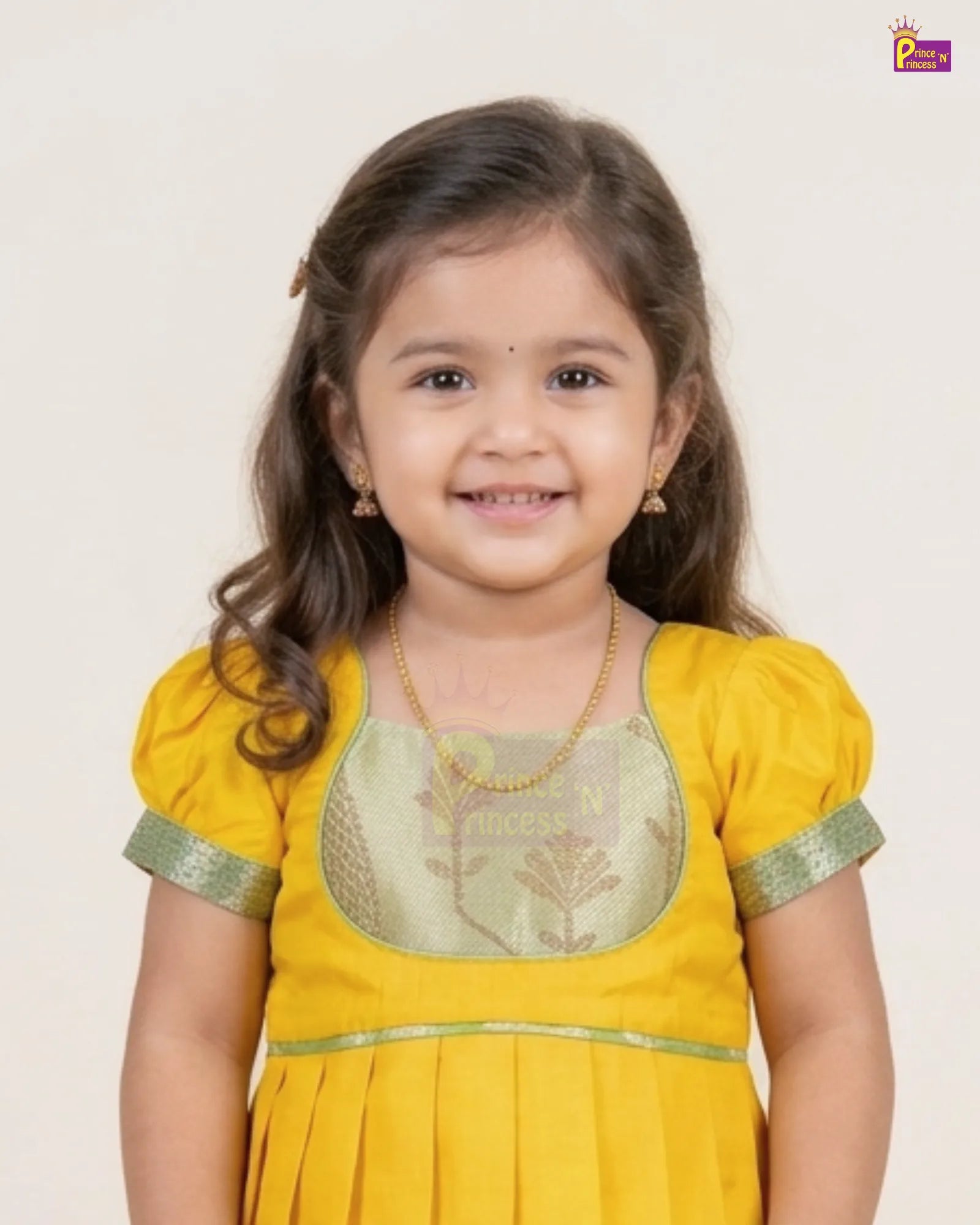 A young girl with long brown hair smiling, wearing a yellow traditional outfit with a light green embroidered chest panel and green trim on the sleeves, set against a plain light background.