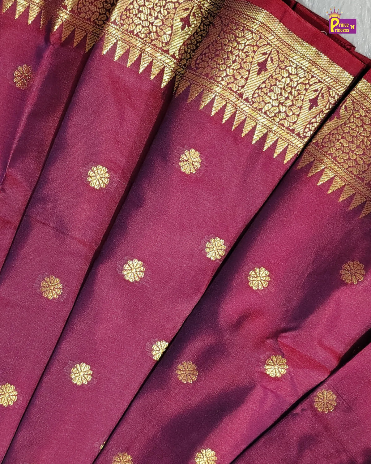 A close-up of a deep magenta silk saree with gold zari woven motifs and a wide decorative gold border.