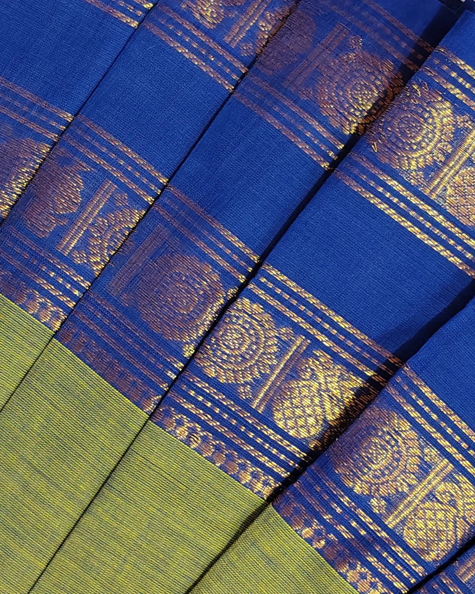 A close-up of a blue and olive green woven fabric, likely a sari, featuring intricate gold zari metallic patterns and borders.