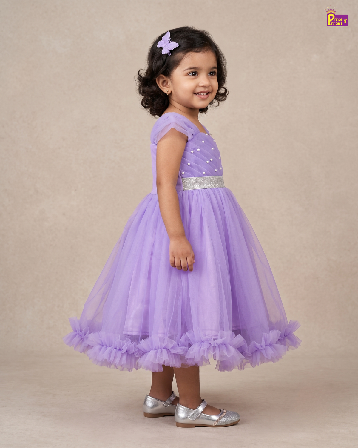 A young girl with dark curly hair wearing a lavender purple party dress with a sparkly bodice, silver belt, and ruffled tulle skirt, standing against a beige studio backdrop.