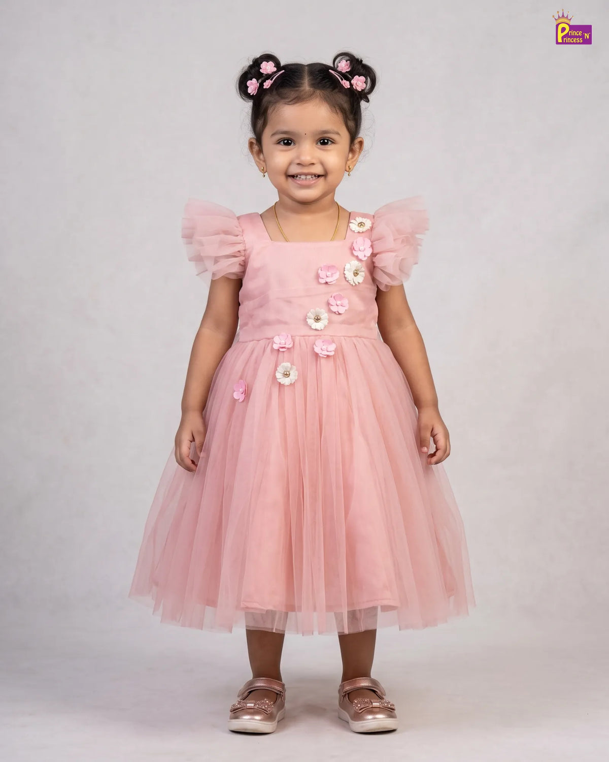 A young girl with dark hair styled in two buns wearing a pink tulle dress with 3D floral embellishments, standing against a light gray studio background.