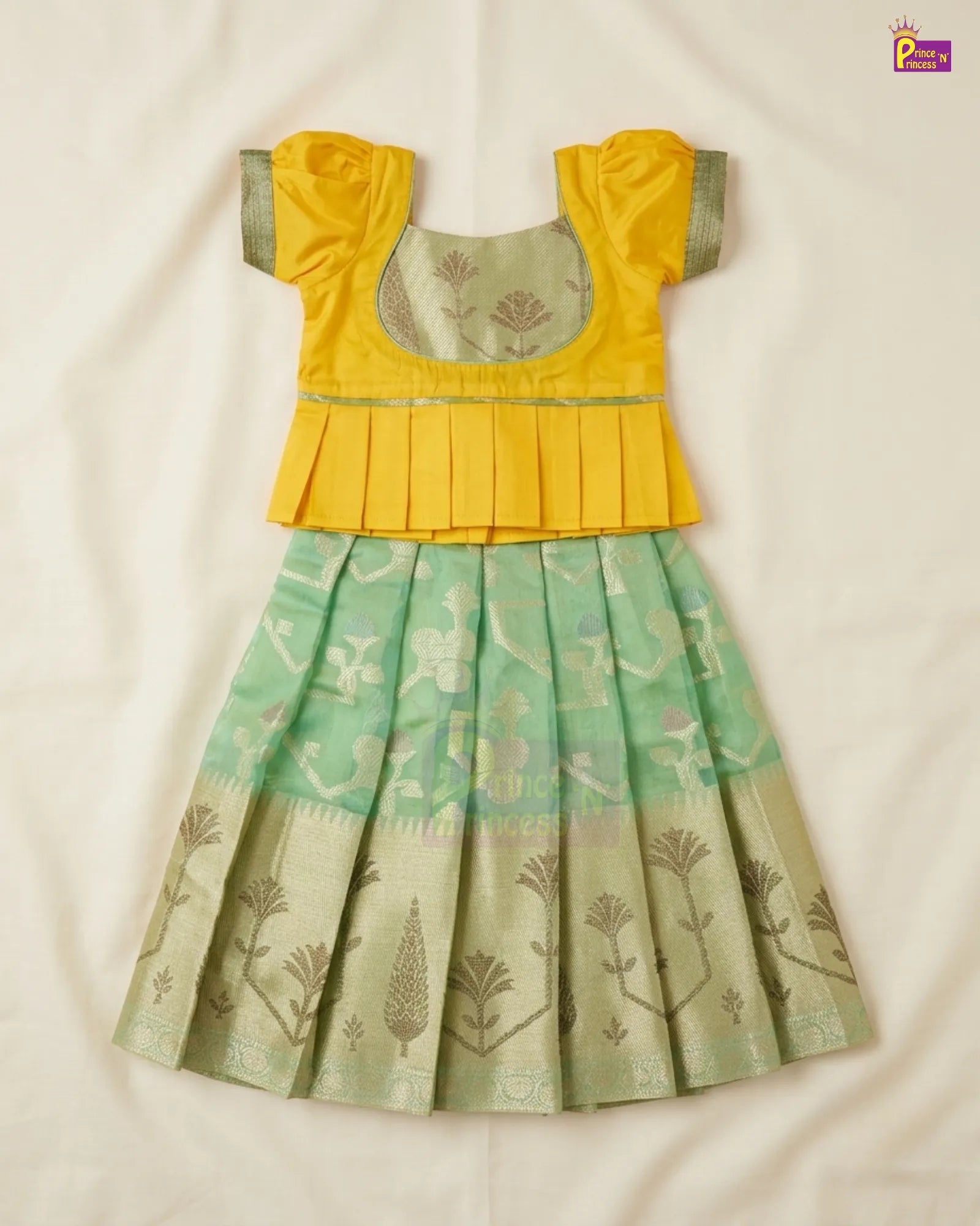 A traditional Indian girl's dress featuring a bright yellow peplum-style top with short puff sleeves and a light green pleated skirt with gold floral motifs, laid flat on a white fabric background.