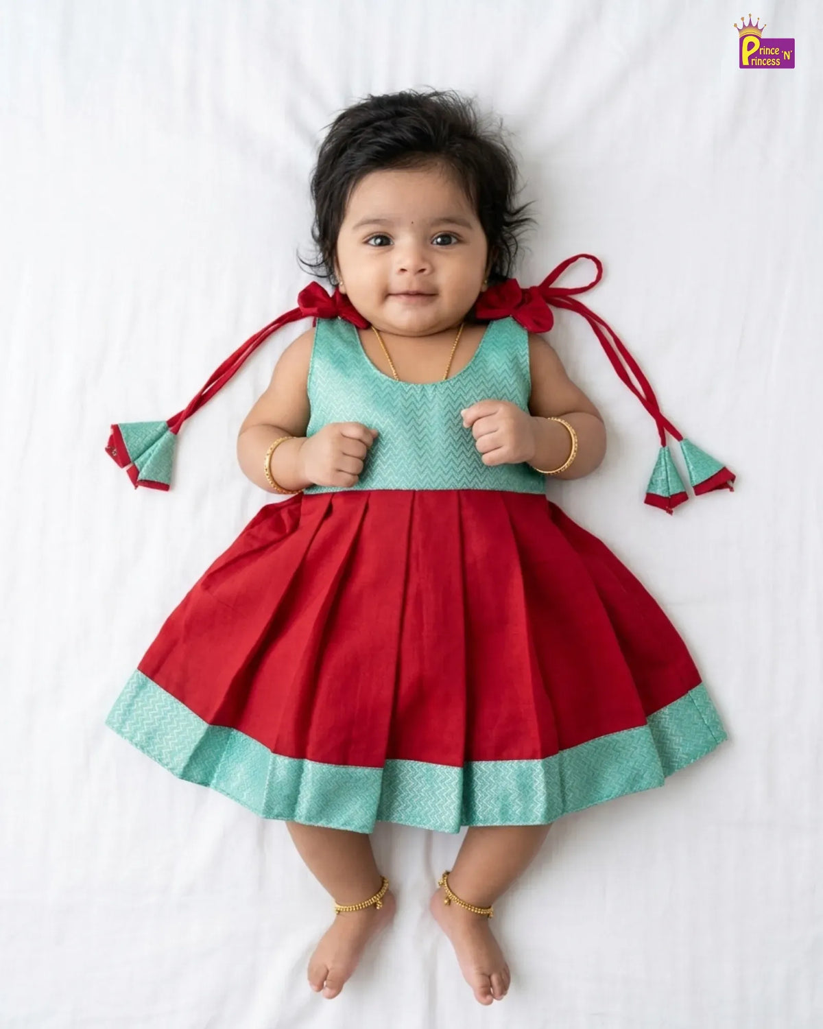 A baby girl lying on a white sheet wearing a red and mint green traditional Indian dress with red ribbon ties on the sides. She is wearing gold bangles and ankle chains.