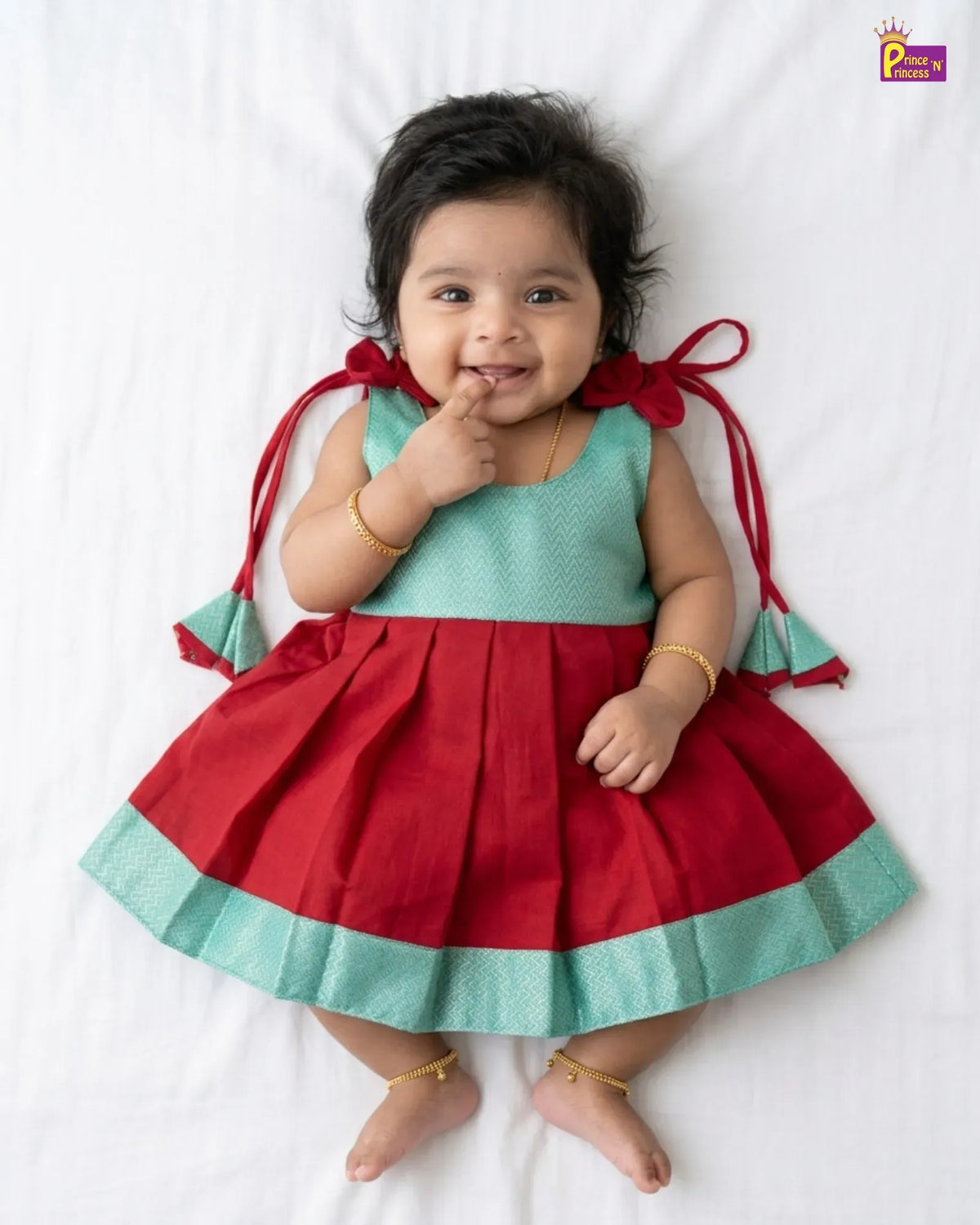A baby girl lying on a white sheet wearing a sleeveless red and mint green dress with red bow-tie details on the shoulders. The brand logo 'Princess P' is visible in the top right corner.