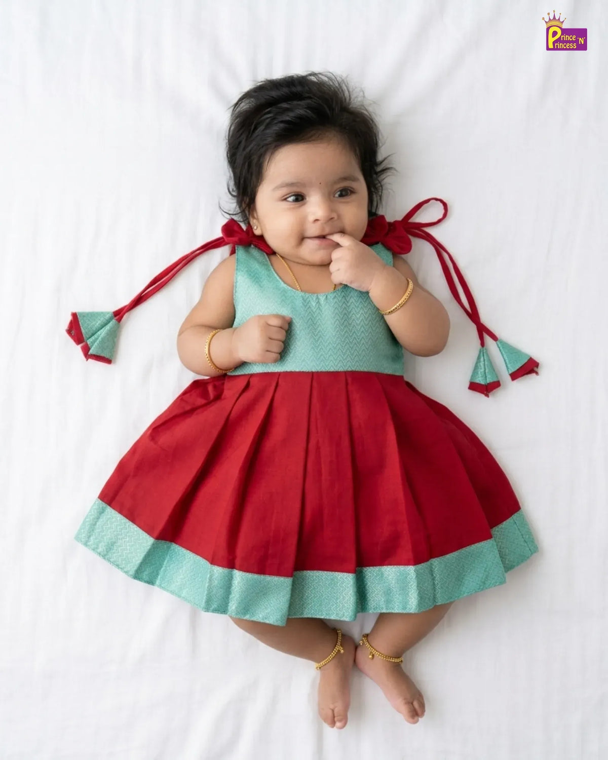A baby girl lying on a white sheet wearing a two-tone red and mint green dress with red ribbon ties on the shoulders. The brand logo 'Princess Padmini' is visible in the top right corner.