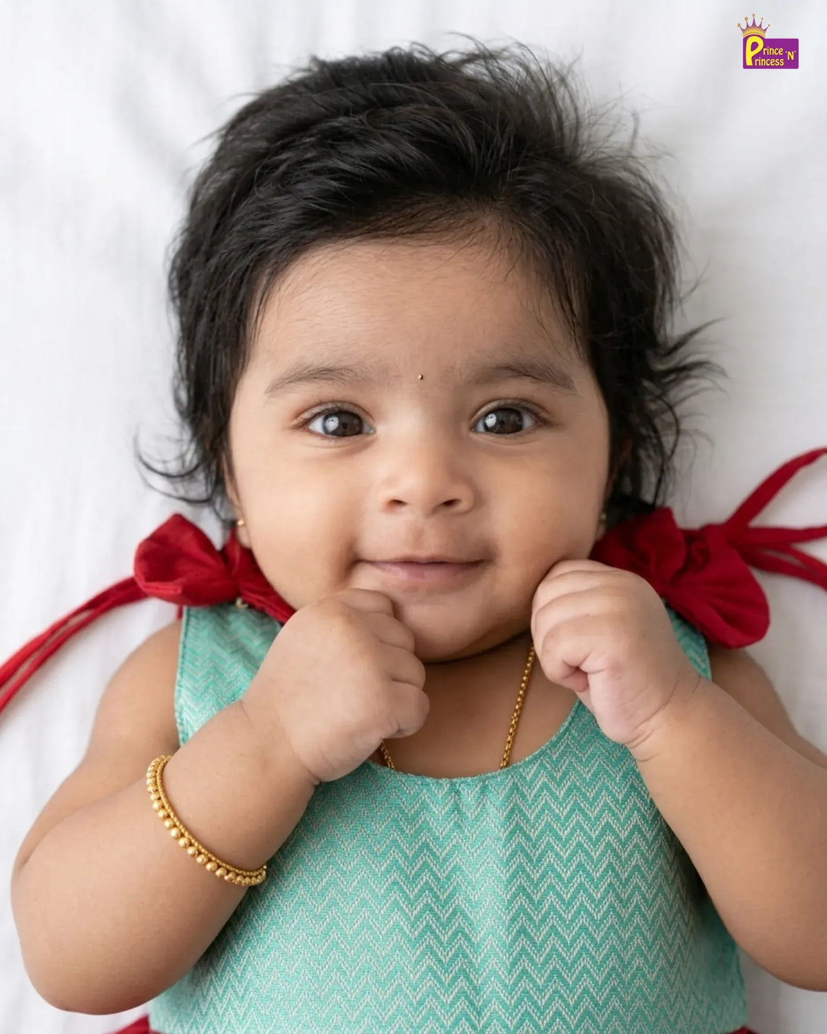 A close-up portrait of a baby lying on a white background, wearing a mint green chevron-patterned sleeveless top with red ribbon accents at the shoulders and a gold chain necklace.