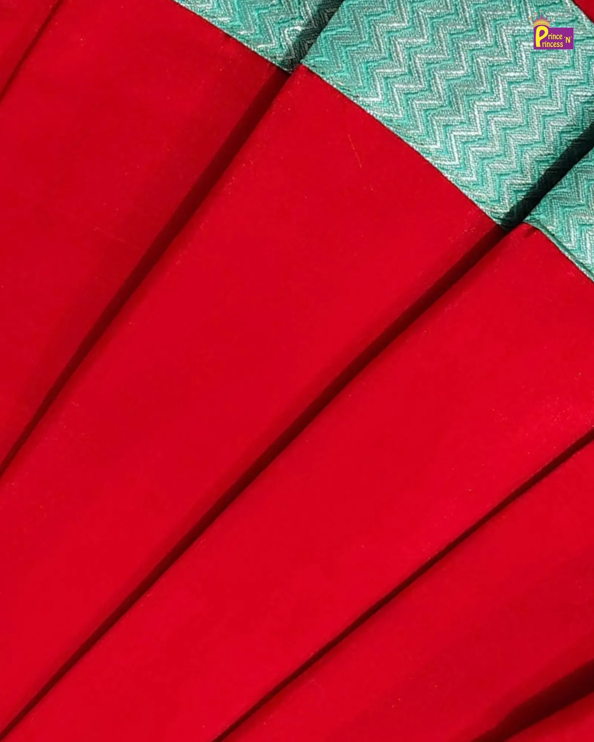 A close-up of a bright red fabric with diagonal pleats and a contrasting teal woven border featuring a white zigzag pattern.