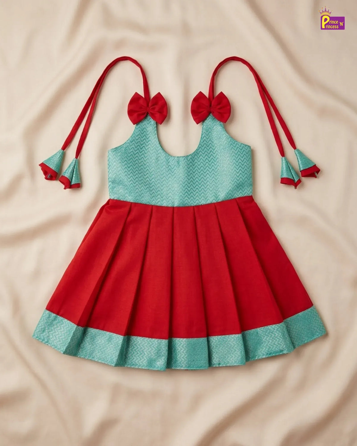 A sleeveless baby girl's dress with a red pleated skirt, a light teal patterned bodice, red shoulder straps with bow and tassel details, and a matching red and teal hem border, laid flat on a beige fabric background.