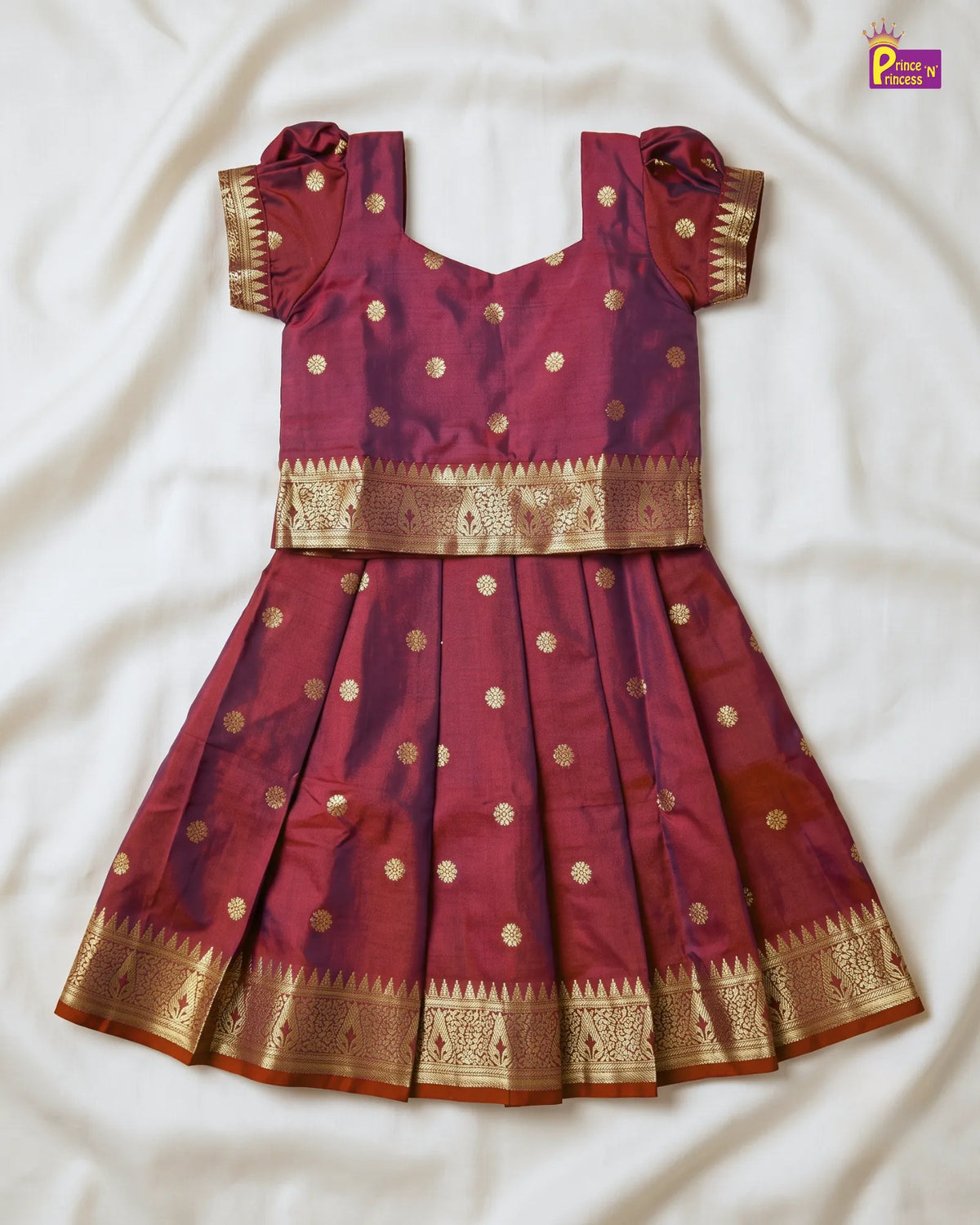 A traditional Indian girl's salwar kameez outfit in maroon with gold zari embroidery, laid flat on a white fabric background. The set includes a sleeveless top with short puff sleeves, a matching pleated skirt, and gold motifs throughout.
