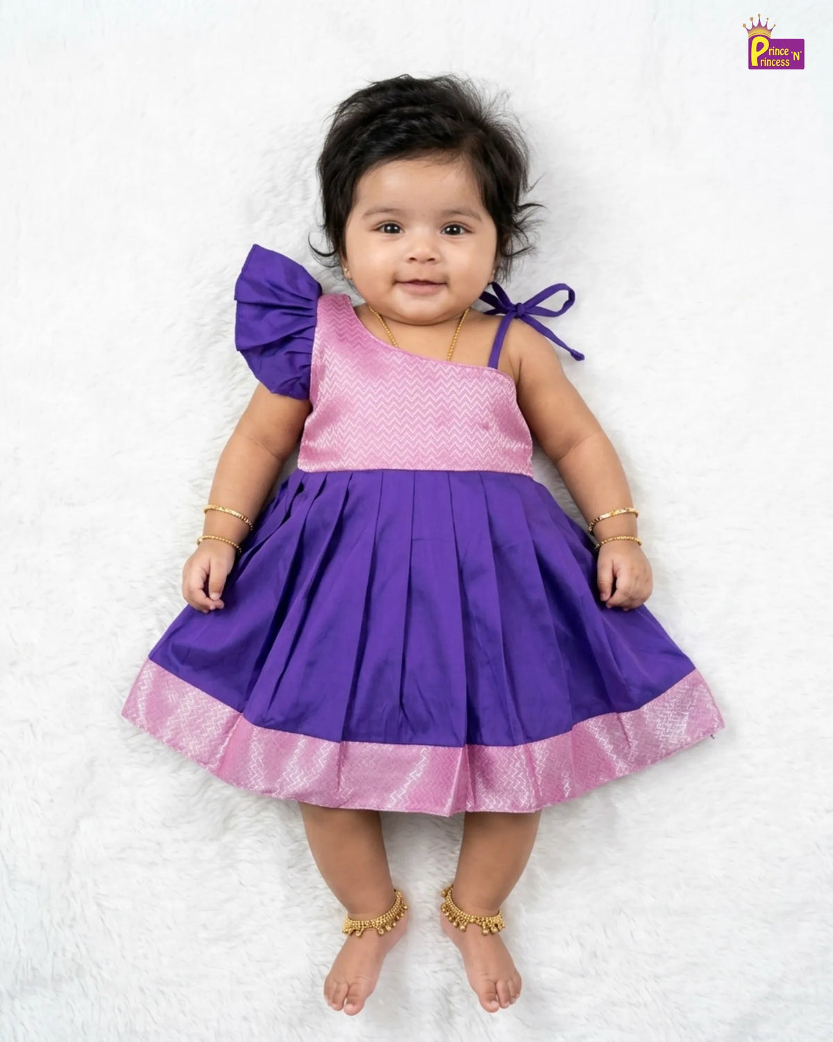 A baby girl wearing a purple and pink dress, lying on a white textured blanket. The dress has a pink patterned bodice, a purple skirt, and a purple bow on the shoulder.
