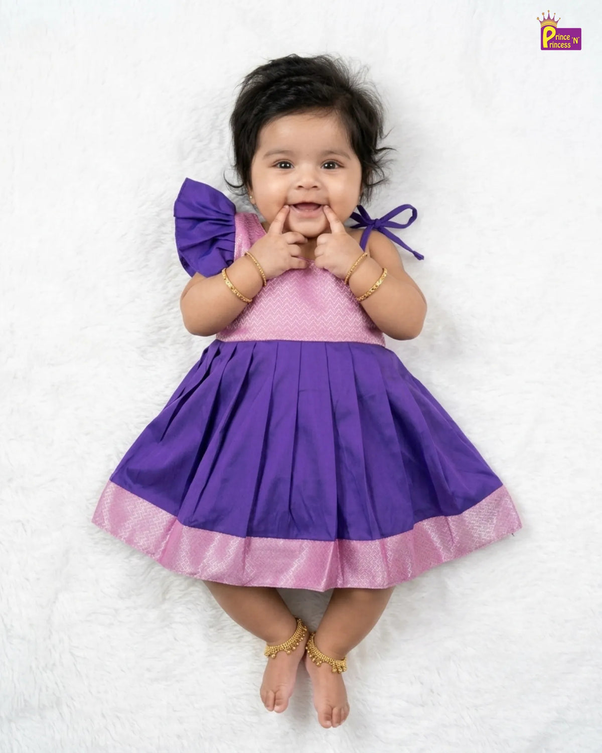 A baby girl lying on a white textured blanket, wearing a purple and pink dress with a pleated skirt, a pink polka-dot bodice, and a purple bow on her shoulder.