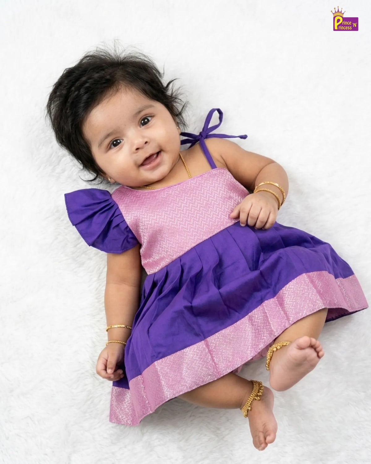 A baby girl wearing a two-tone purple and pink dress, lying on a white textured blanket. The dress has a pink upper bodice with a subtle pattern, a solid purple ruffled shoulder, and a purple skirt with a pink border.