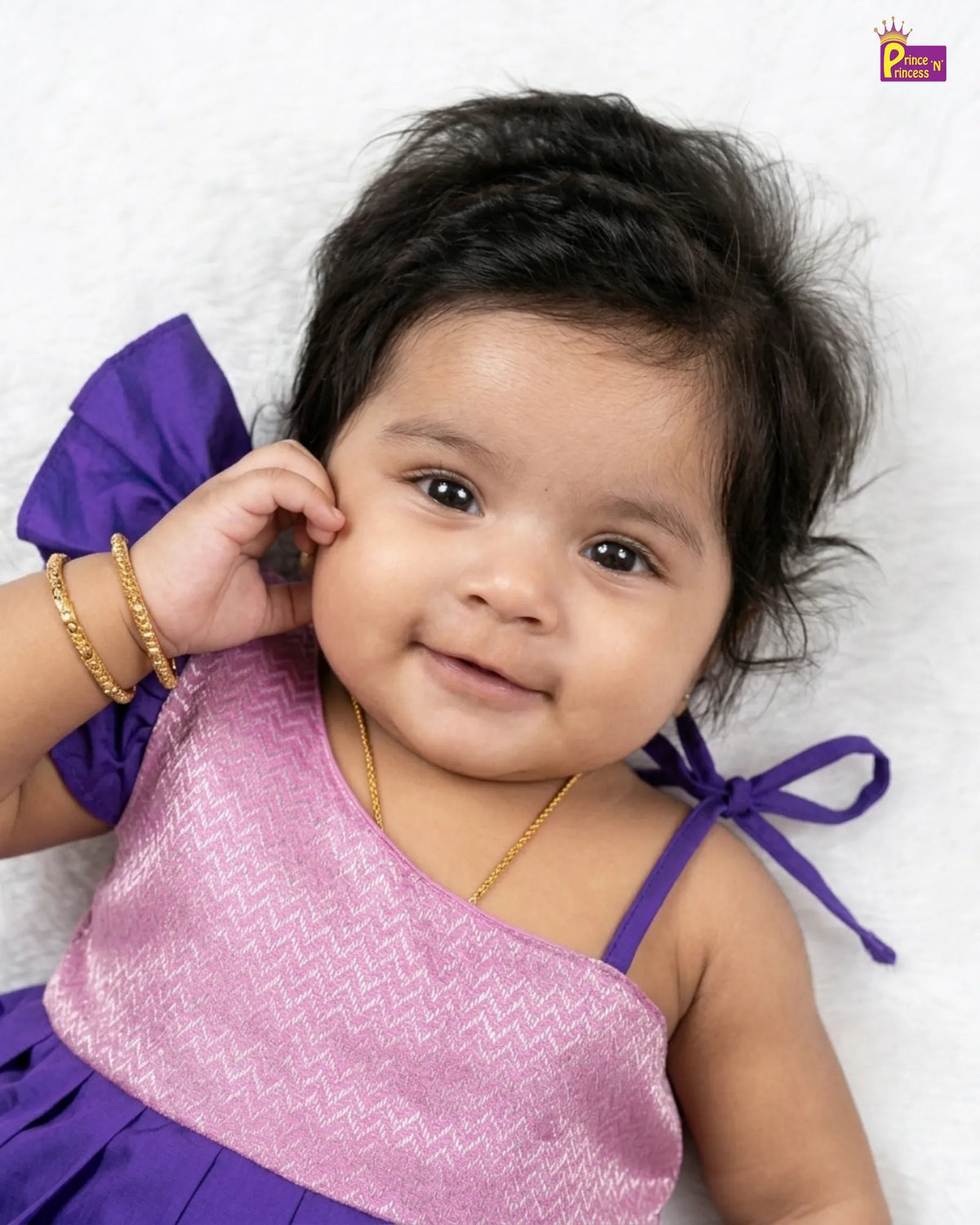 A smiling baby girl wearing a purple and pink dress, lying on a white textured blanket. She has gold bangles and a gold necklace on.