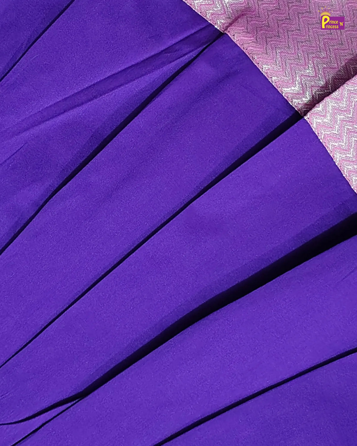 A close-up of a purple fabric with diagonal pleats, showing a contrasting pink and white patterned border in the top right corner.