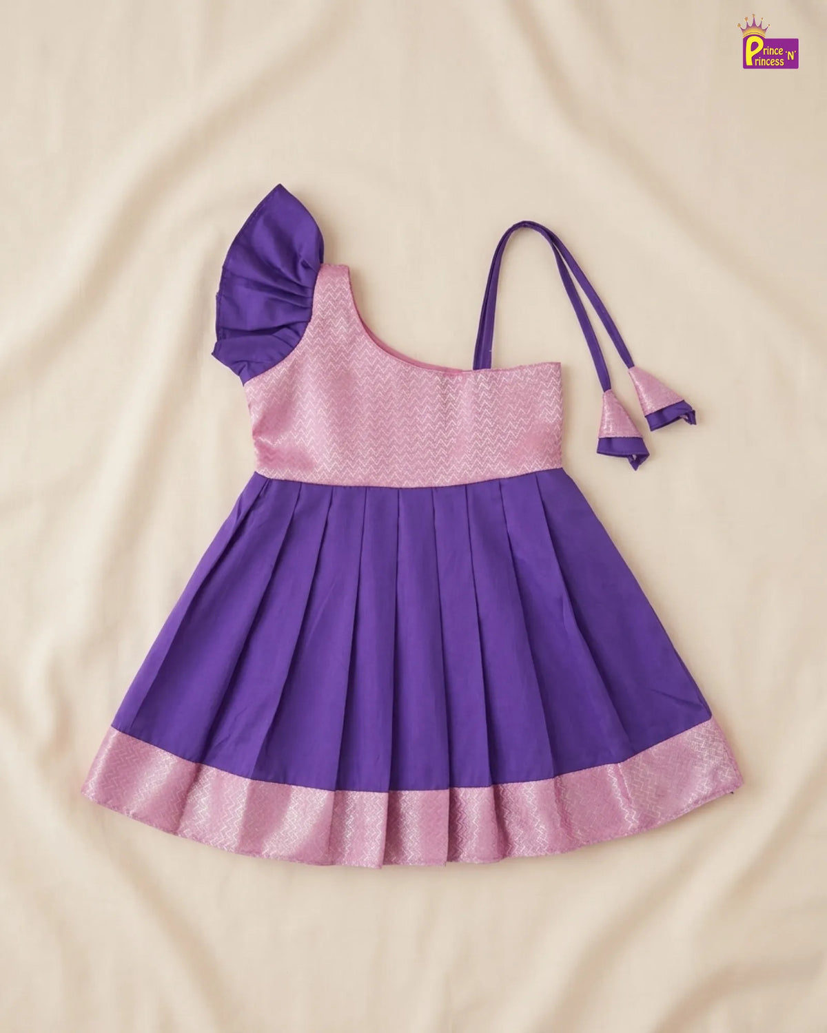 A purple and pink toddler girl's dress laid flat on a beige fabric background. The dress features a solid purple pleated skirt, a light pink textured bodice, a ruffled shoulder strap, and a matching pink and purple tie detail on the side.