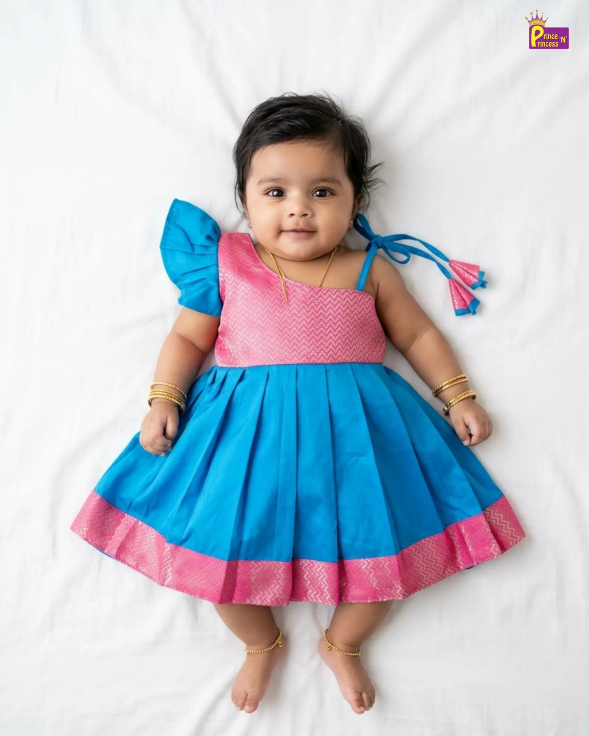 A baby girl lying on white sheets wearing a bright blue and pink traditional Indian dress with a pleated skirt, pink patterned bodice, and a blue ruffled shoulder tie.