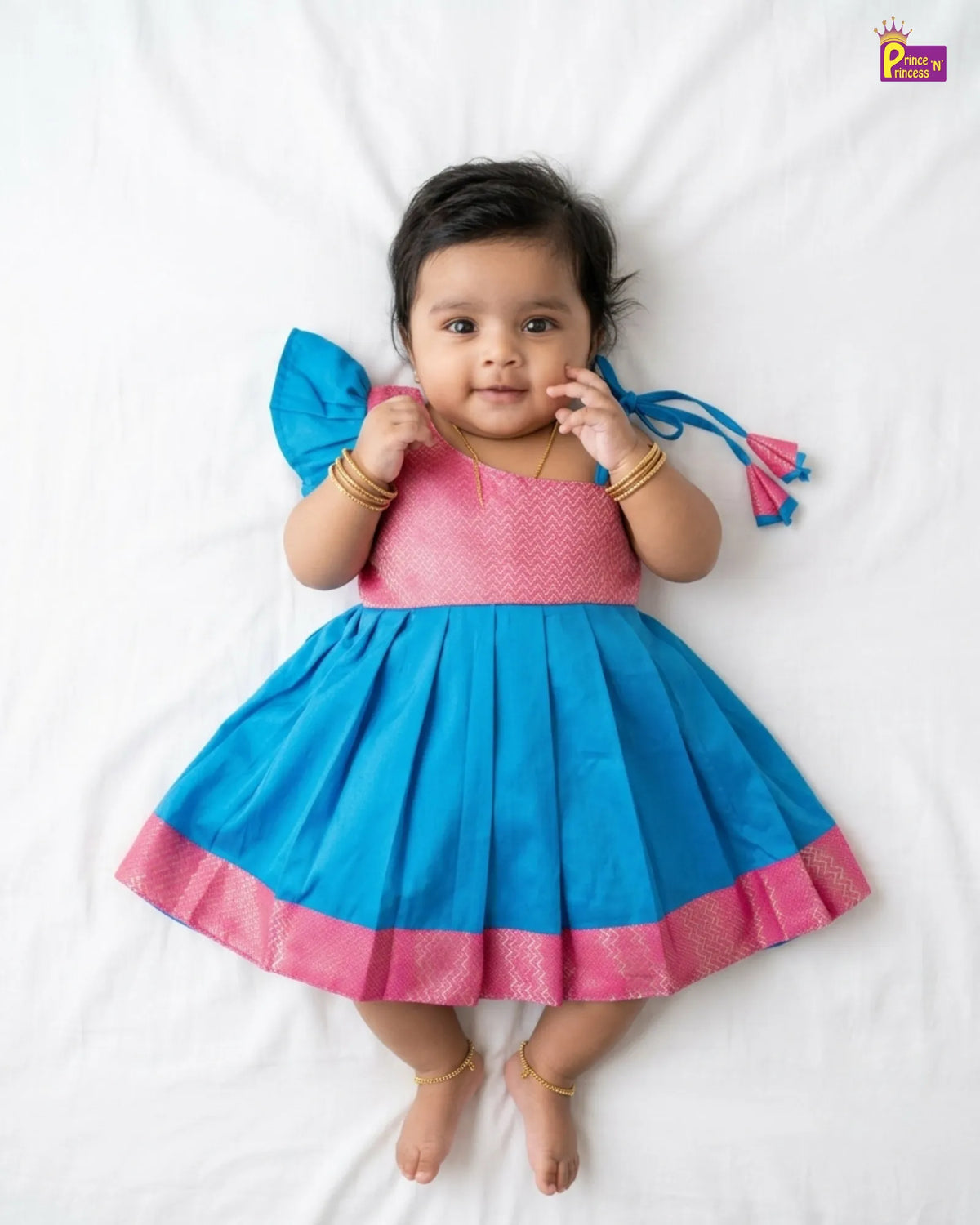A baby girl lying on a white sheet wearing a bright blue and pink traditional Indian dress with gold bangles and anklets.
