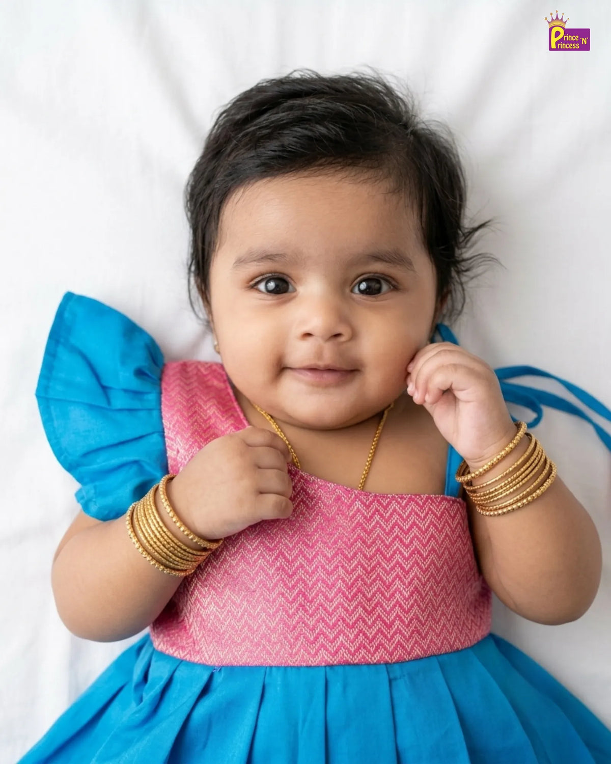 A baby girl wearing a traditional Indian dress with a pink chevron-patterned bodice and a bright blue flared skirt, lying on a white background. She is wearing gold bangles and a gold necklace.