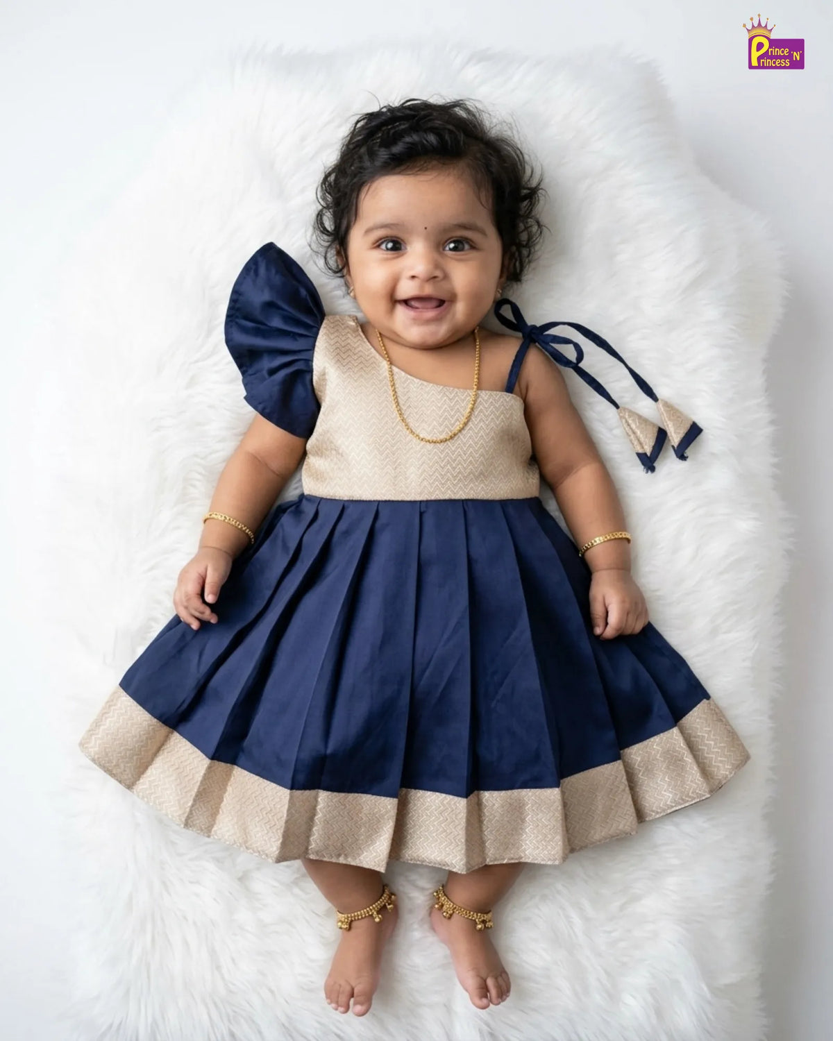 A baby girl wearing a navy blue and beige traditional Indian dress, lying on a white faux fur rug. The dress features a beige bodice with a subtle gold pattern, a navy blue pleated skirt with a beige border, and a large navy blue ruffled shoulder detail.