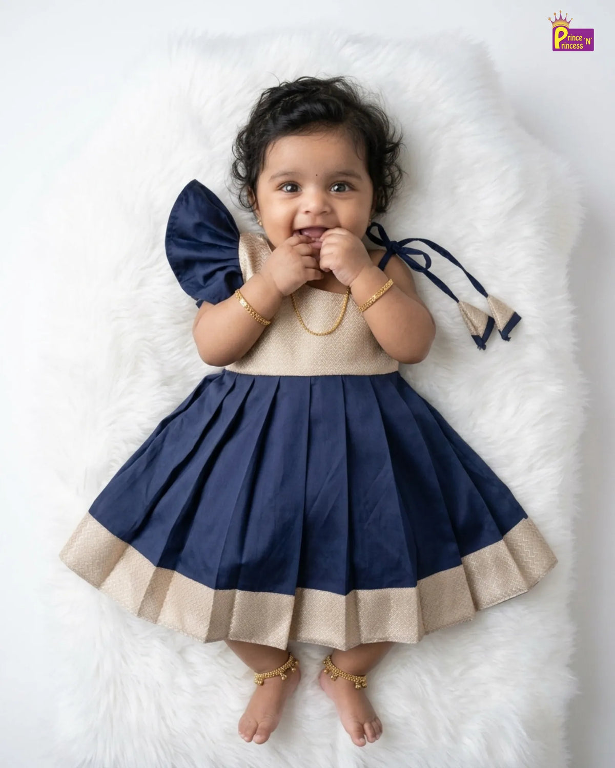 A baby girl wearing a navy blue and beige pleated dress with a ruffled hem and a matching navy bow on the shoulder, lying on a white faux fur rug.
