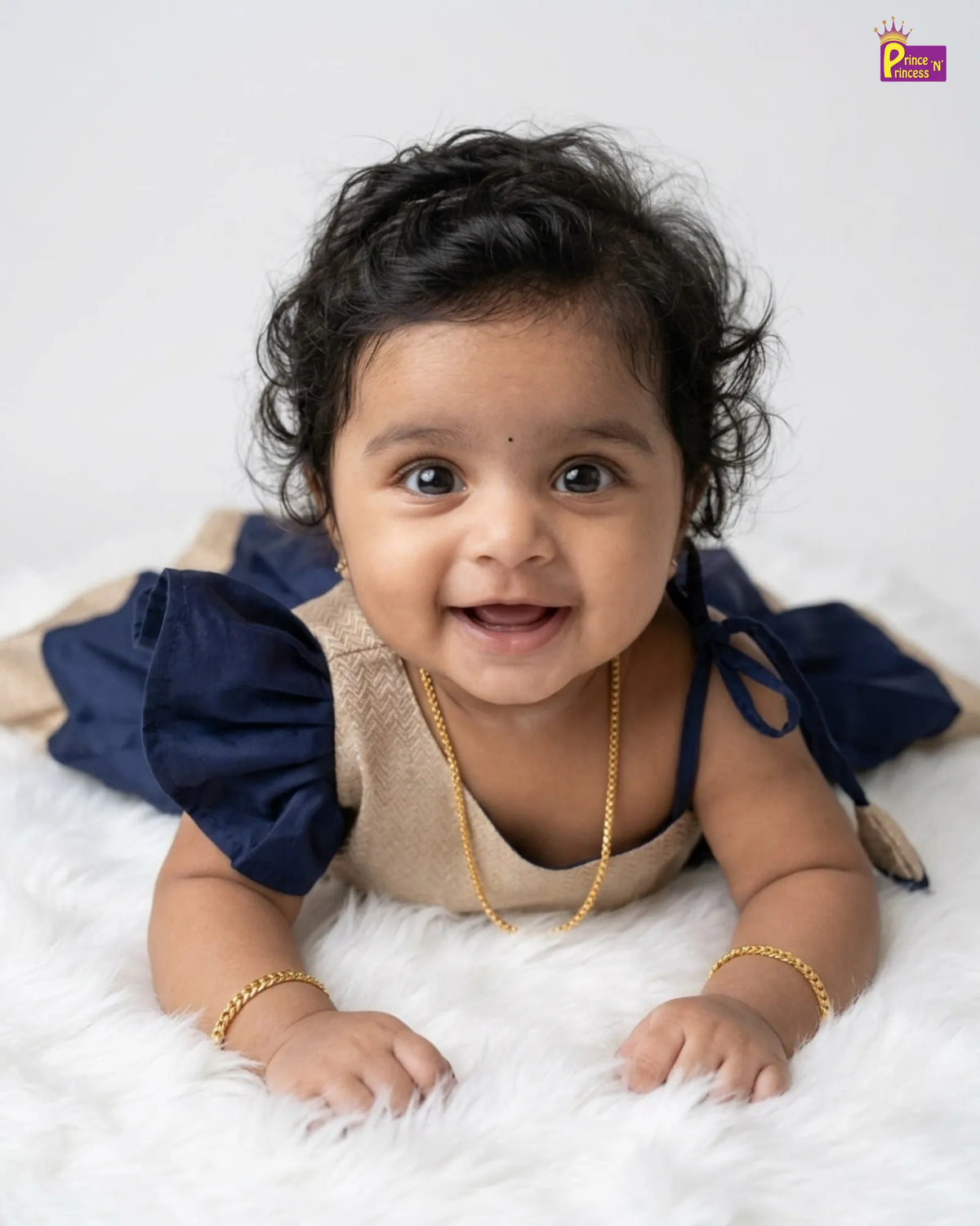 A smiling baby girl lying on her stomach on a white faux fur rug, wearing a beige and navy blue dress with gold jewelry. A 'P.
