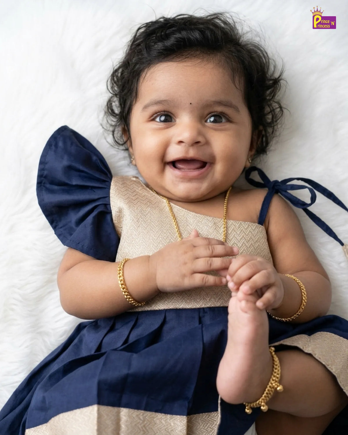 A smiling toddler girl lying on a white textured blanket, wearing a navy blue and beige dress with gold jewelry.