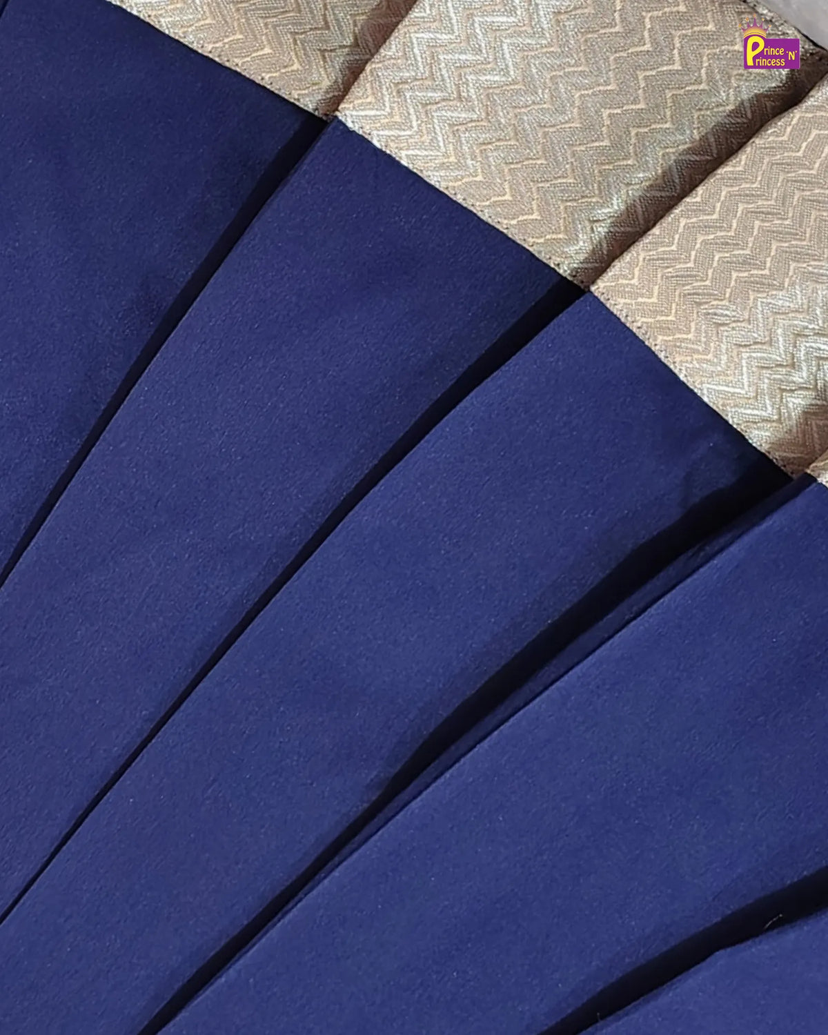 A close-up of a pleated navy blue fabric garment, likely a sari, with a contrasting woven beige and gold chevron-patterned border.