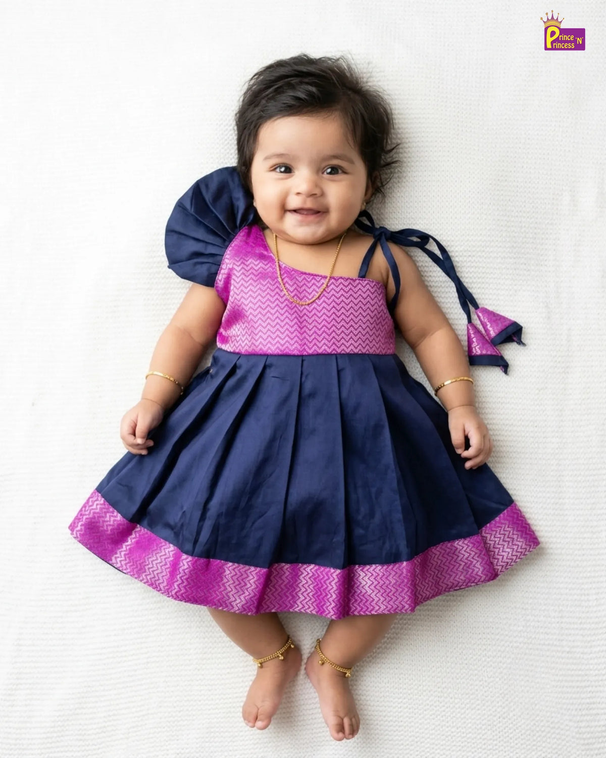 A baby girl wearing a navy blue and magenta traditional dress, lying on a white textured blanket. The dress features a pleated skirt, a magenta patterned bodice, and a large navy blue shoulder bow.