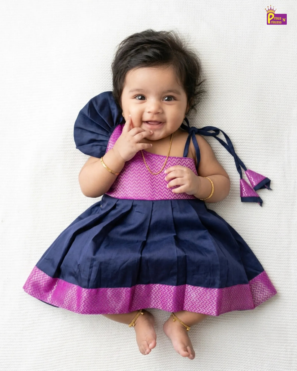A baby girl lying on a white textured blanket, wearing a navy blue and magenta traditional Indian dress with gold jewelry.