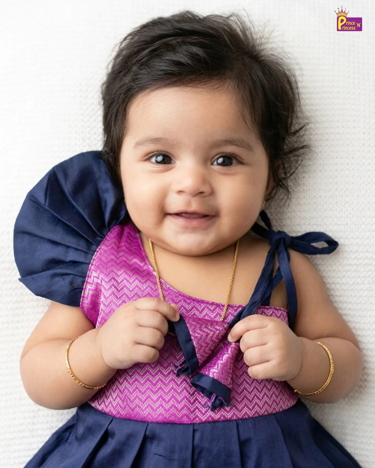 A baby girl wearing a purple and navy blue dress, lying on a white textured blanket. The dress features a purple chevron-patterned bodice with a pink zigzag overlay, navy blue puffed shoulder details, and navy blue tie straps.