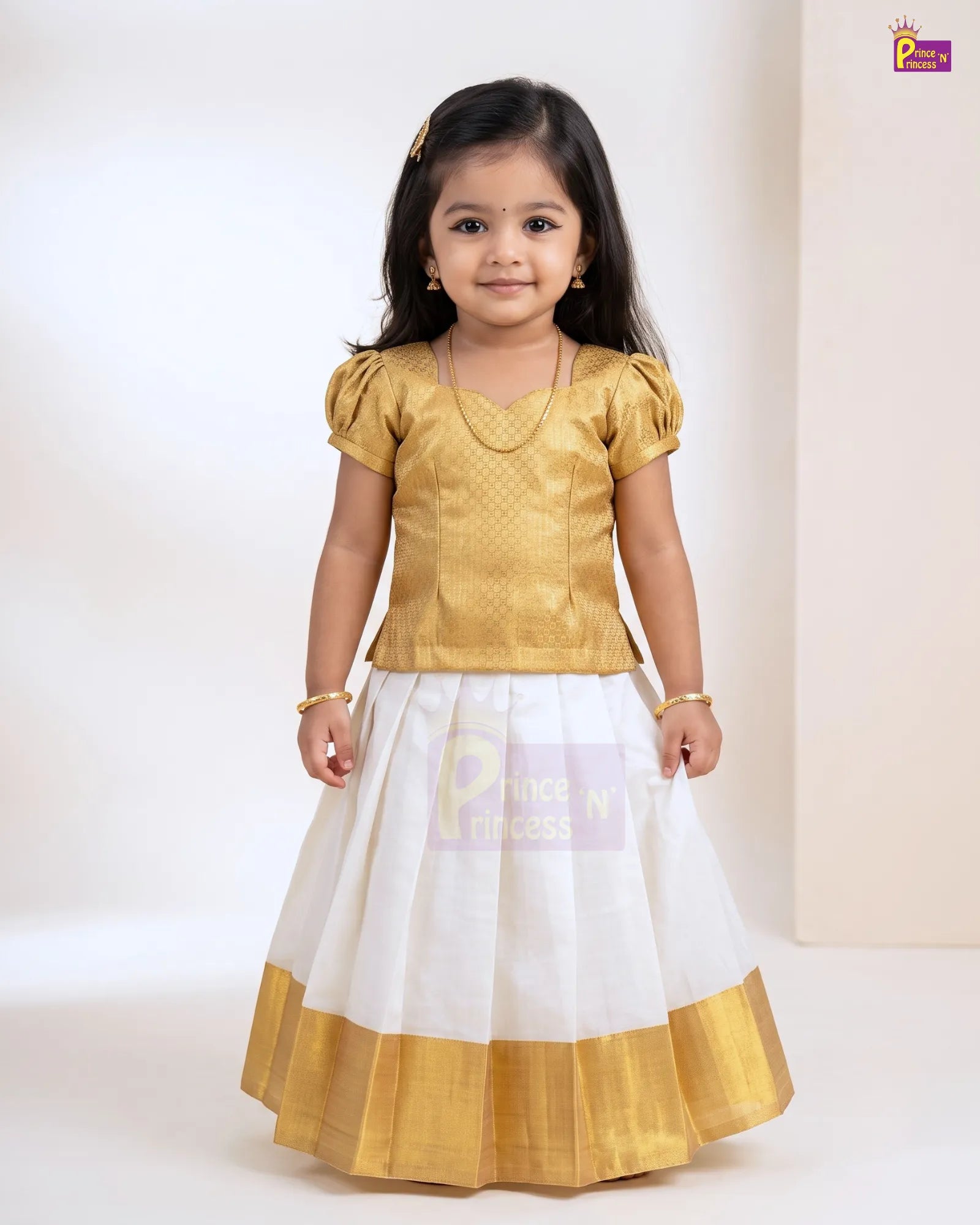 A young girl modeling a traditional Indian outfit consisting of a gold metallic short-sleeve blouse and a white skirt with a gold border, set against a plain light beige studio background.