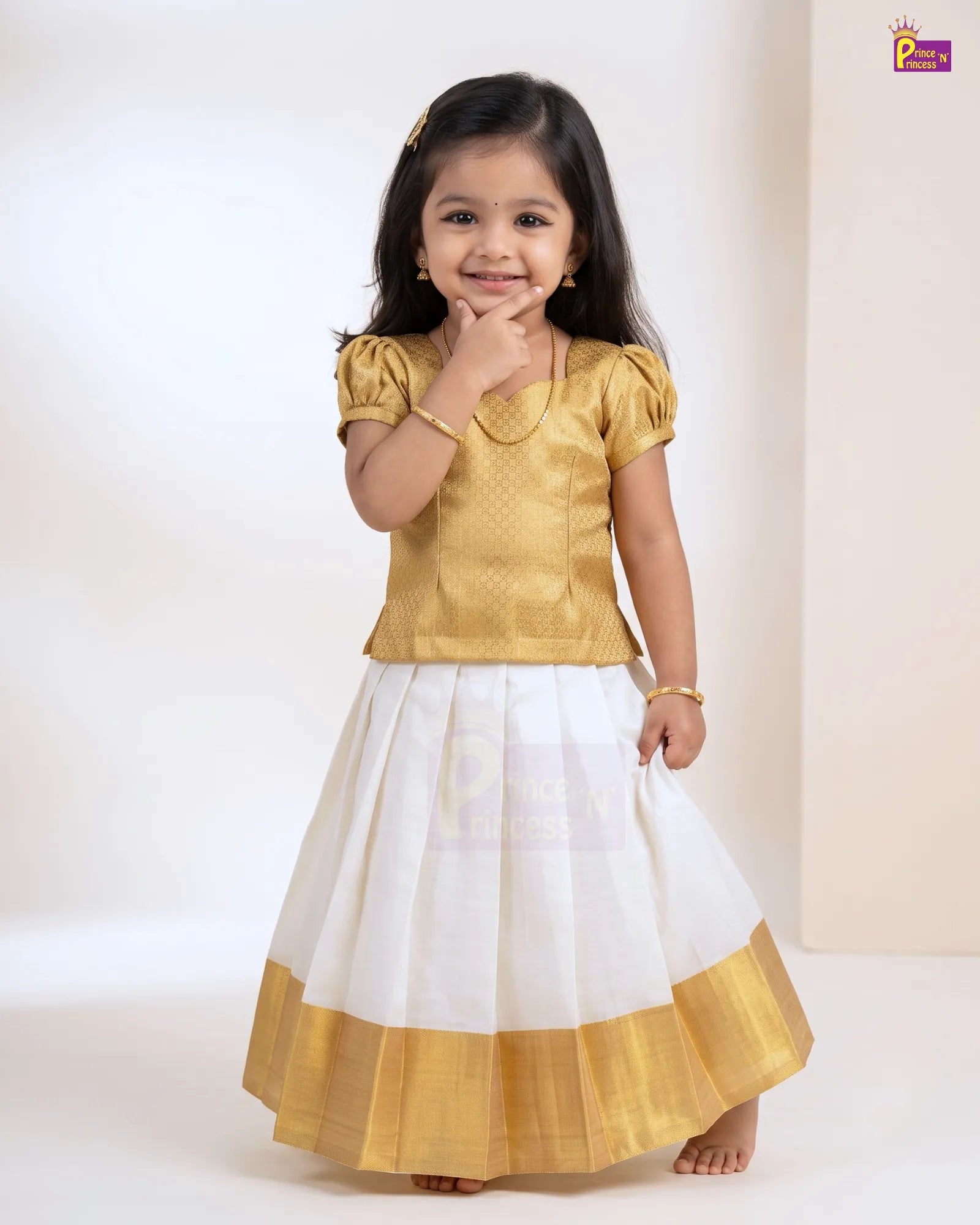 A young girl modeling a traditional Indian dress consisting of a gold short-sleeve top and a white skirt with a gold border, set against a plain light beige studio background.