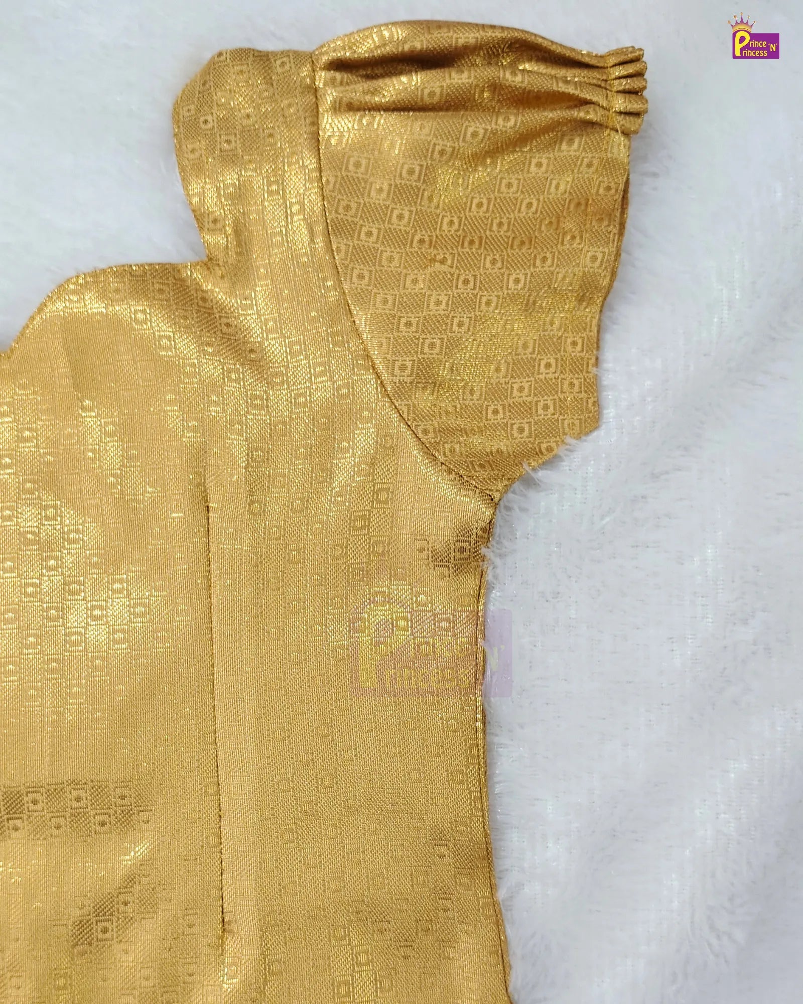 A close-up of a gold-colored fabric garment, likely a traditional dress or sari, featuring a textured woven pattern and a gathered sleeve, laid flat on a white faux fur surface.
