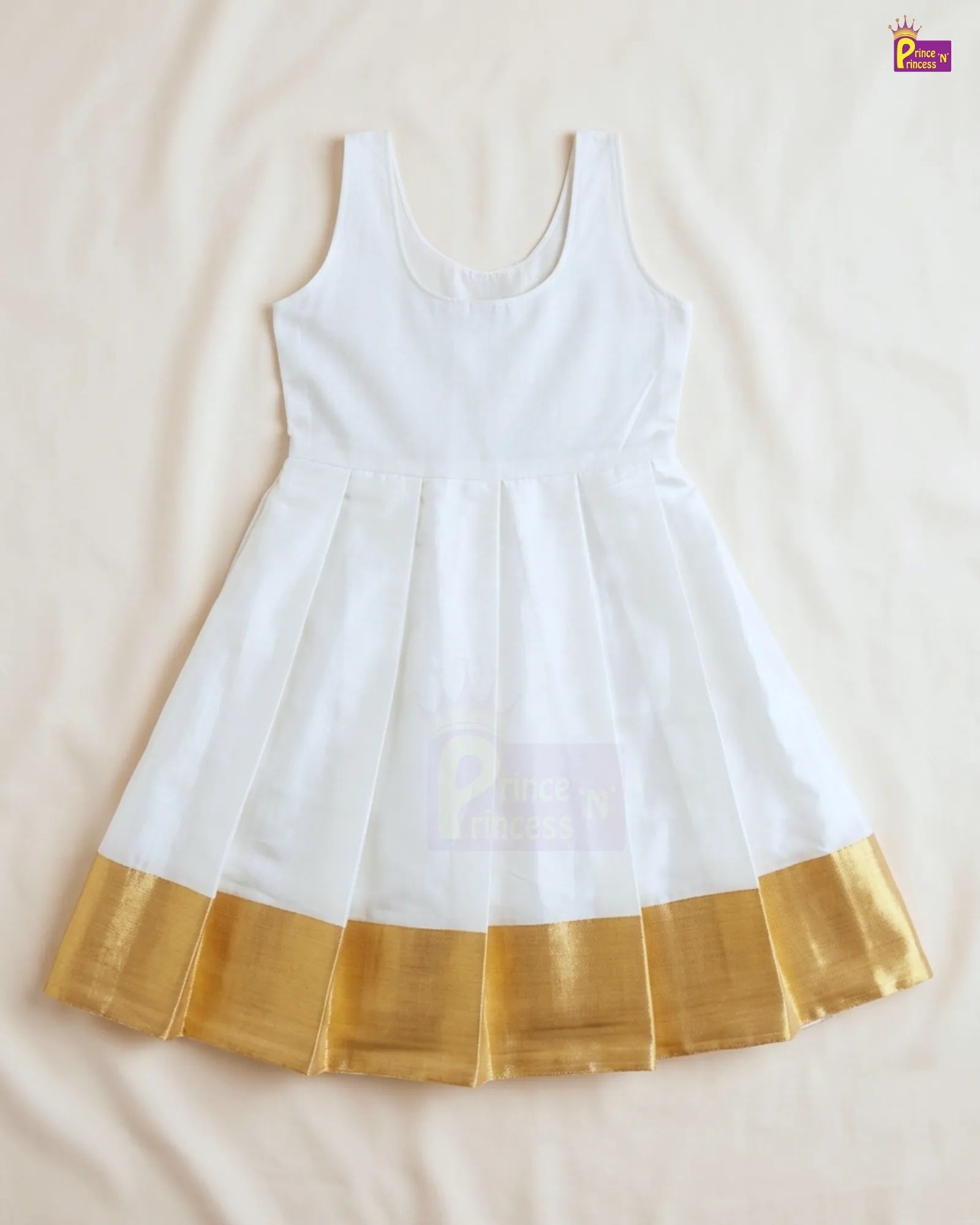 A white sleeveless children's dress with a pleated skirt and a shiny gold border along the hem, laid flat on a light beige fabric background.
