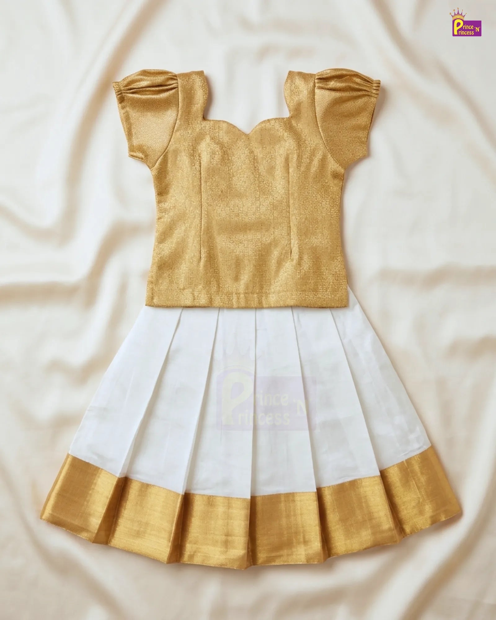 A traditional Indian girl's dress set featuring a metallic gold blouse with puffed sleeves and a white pleated skirt with a gold border, laid flat on a cream-colored fabric background.