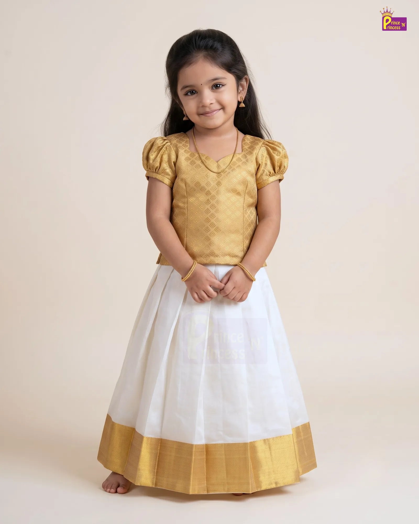 A young girl modeling a traditional Indian dress with a gold metallic blouse and a white skirt featuring a gold border, set against a plain beige studio background.