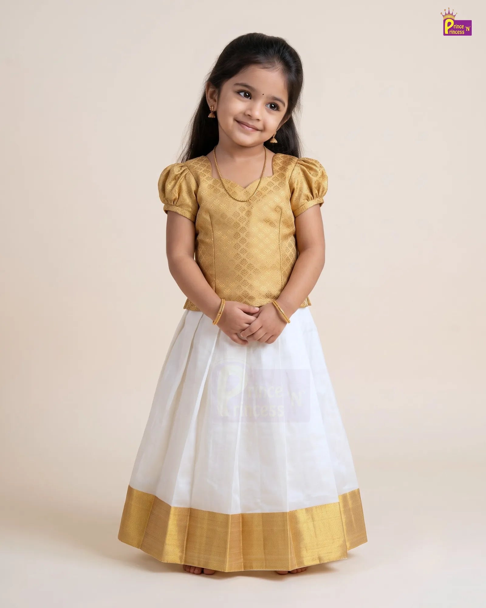 A young girl modeling a traditional Indian dress with a gold textured blouse and a white skirt featuring a gold border, set against a plain beige studio background.