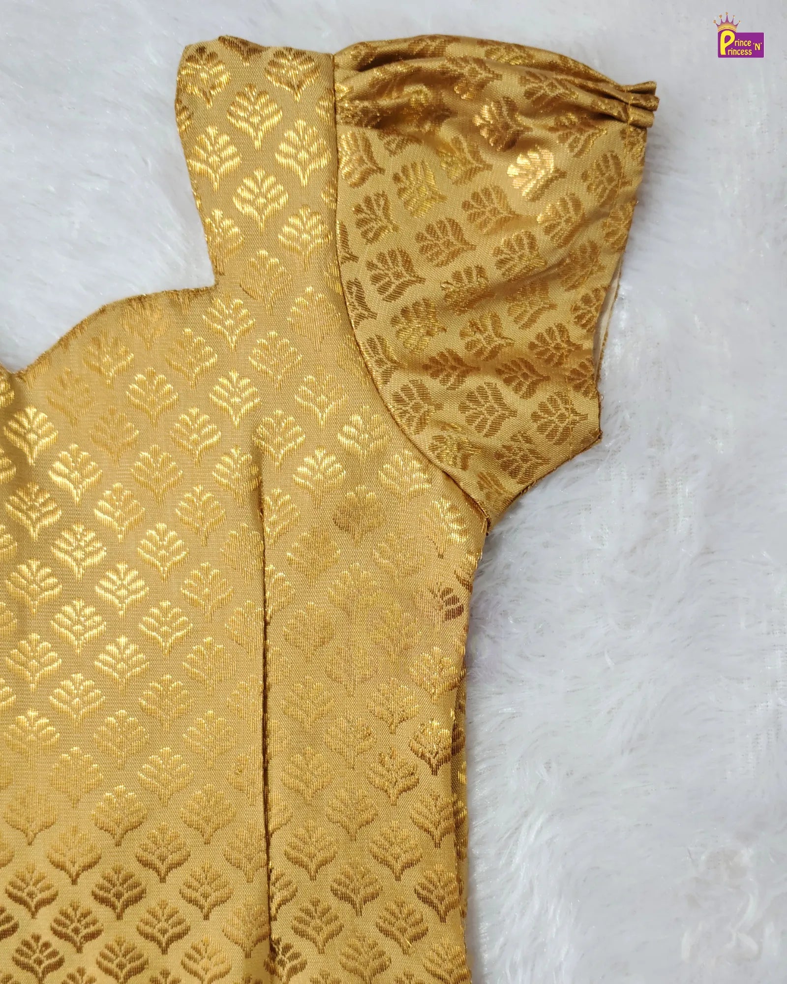A close-up of a gold brocade fabric garment or textile piece laid flat on a white faux-fur surface, showing a repeating leaf-like woven pattern and gathered ruffle detail.