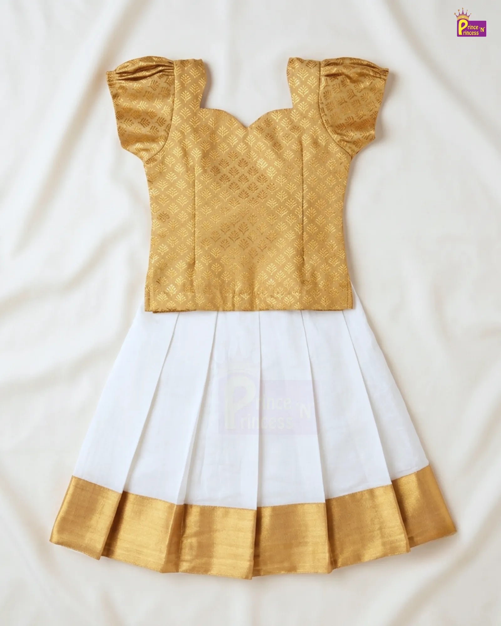 A traditional Indian toddler girl's dress consisting of a gold metallic brocade blouse with puffed sleeves and a white pleated skirt with a gold border, laid flat on a white fabric background.