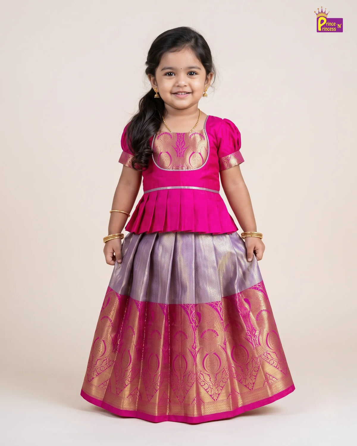 A young girl modeling a traditional Indian ethnic outfit consisting of a bright pink peplum top and a matching flared skirt with purple and gold zari embroidery, set against a plain beige background.