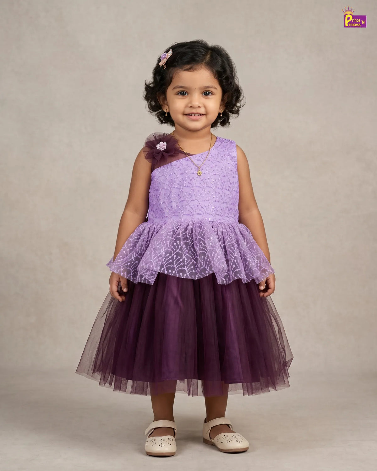 A young girl with dark curly hair wearing a two-tone purple dress, standing against a neutral beige studio background. The dress features a light purple lace one-shoulder top with a ruffled overlay and a darker purple tulle skirt.