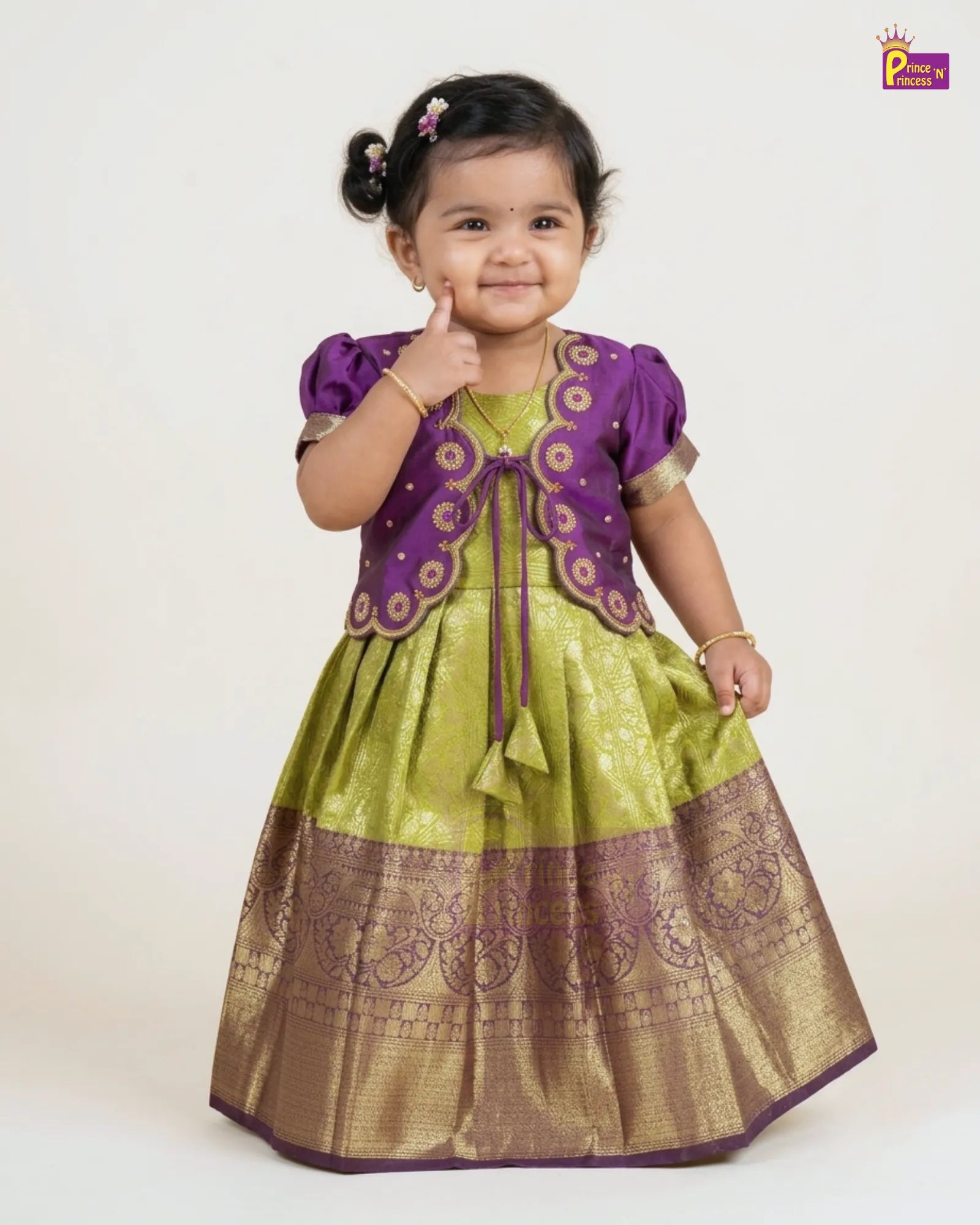 A young toddler girl modeling a traditional Indian ethnic dress with a purple embroidered vest and a green and gold skirt, posing against a plain off-white background.