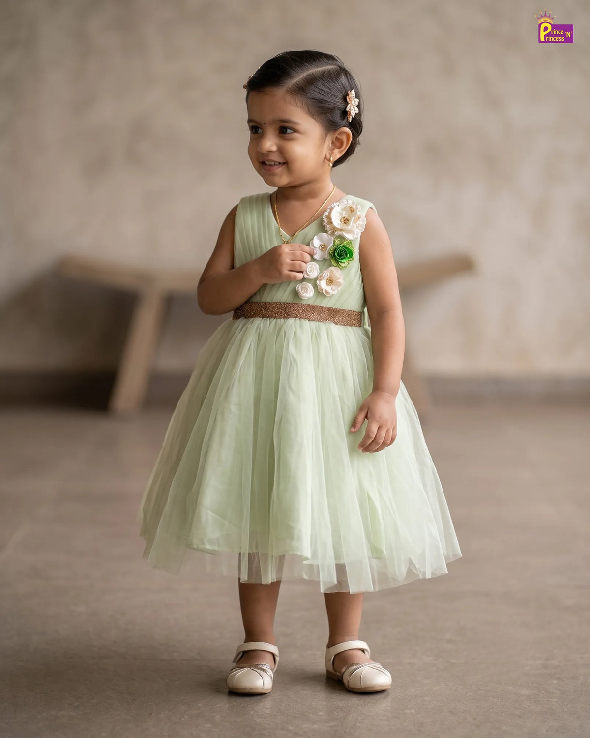 A toddler girl wearing a sleeveless mint green tulle dress with a brown waist belt and 3D floral embellishments on the bodice, standing in a studio setting.