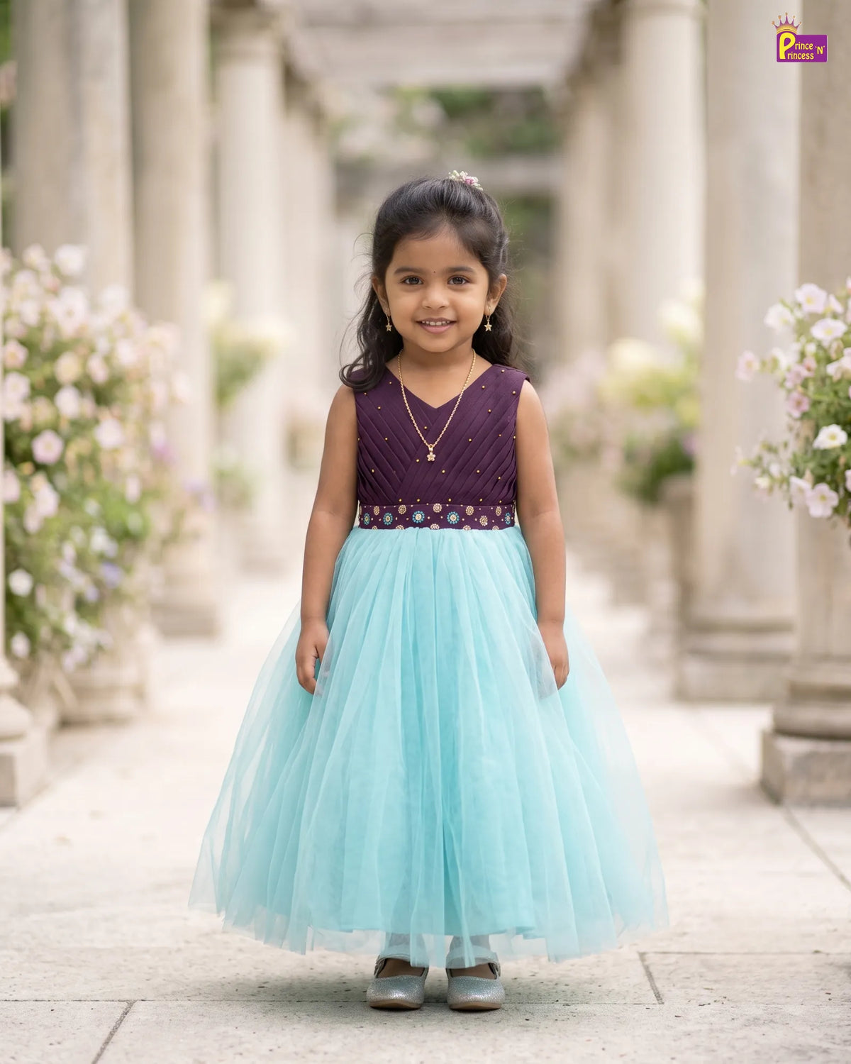 A young girl stands in a stone walkway lined with columns and flowers, wearing a sleeveless formal dress with a deep purple embellished bodice and a voluminous light aqua tulle skirt.